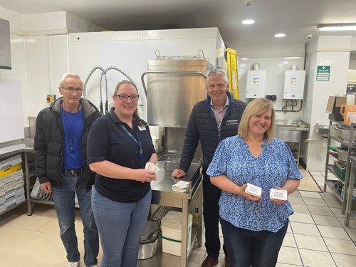Four people in a factory kitchen holding donated food packs