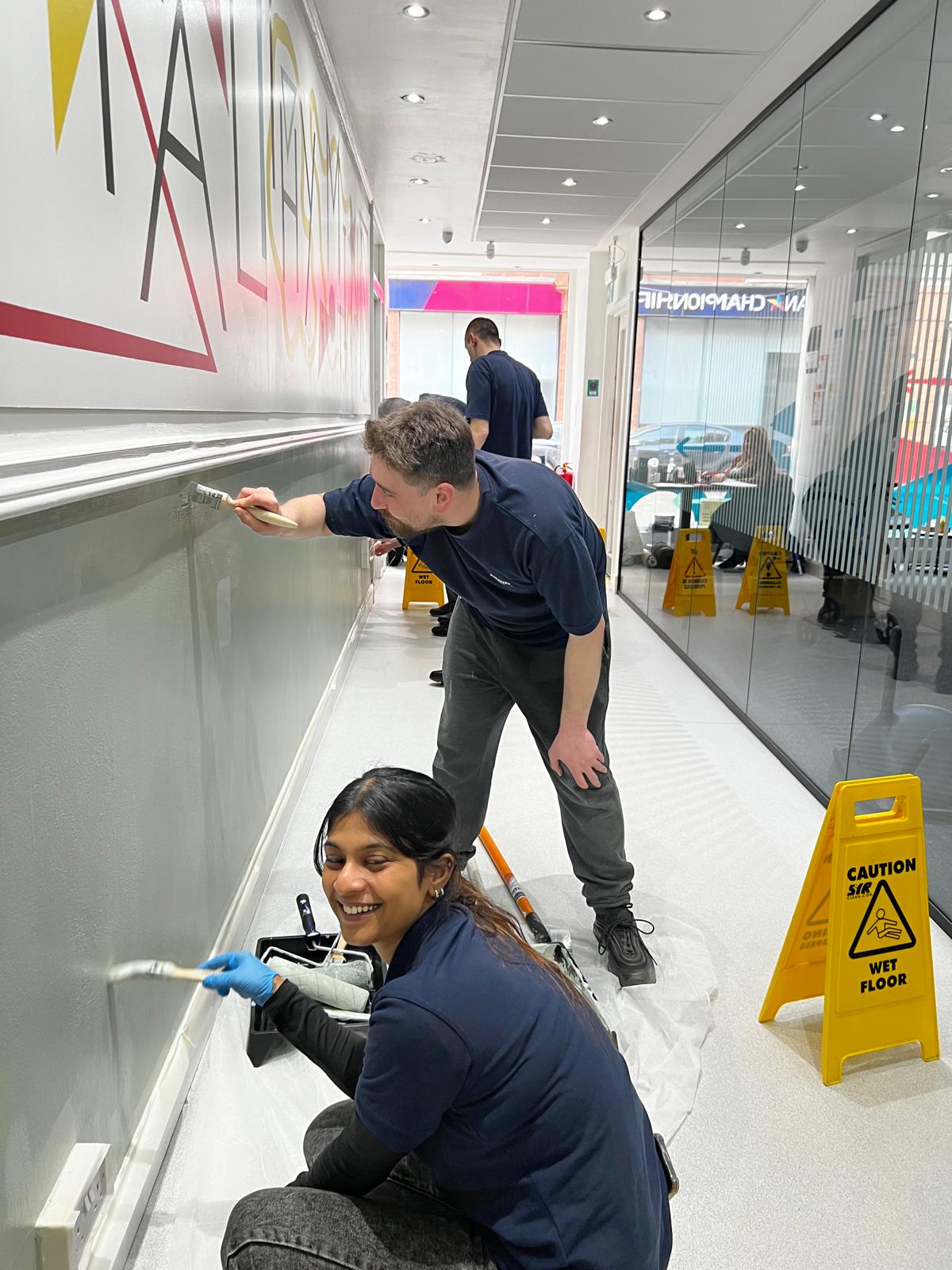 Two Kerry volunteers painting a corridor wall below a mural